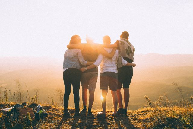 Four people with hands wrapped around each others shoulders while looking at a sunset.