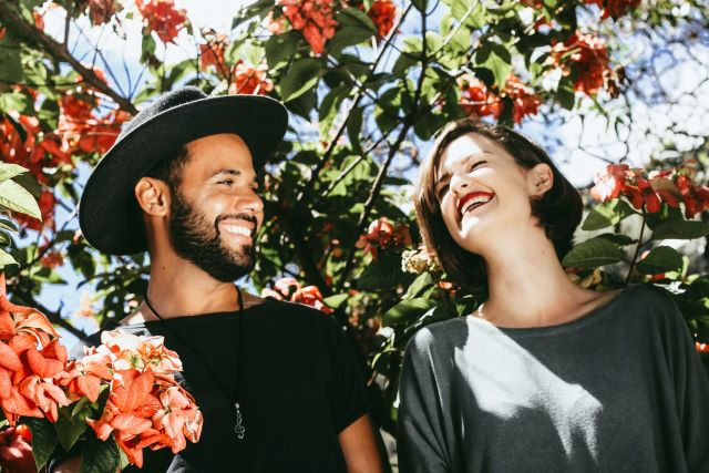 Two people smiling in the sun with a tree filled with flowers behind them.
